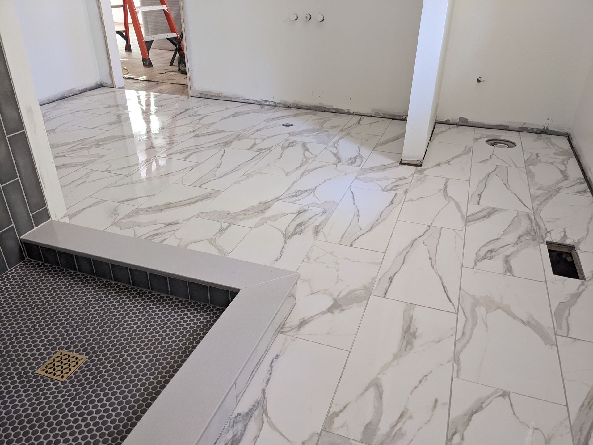 Bathroom under renovation with white marble-patterned floor tiles being installed. Gray border surrounds the shower area.