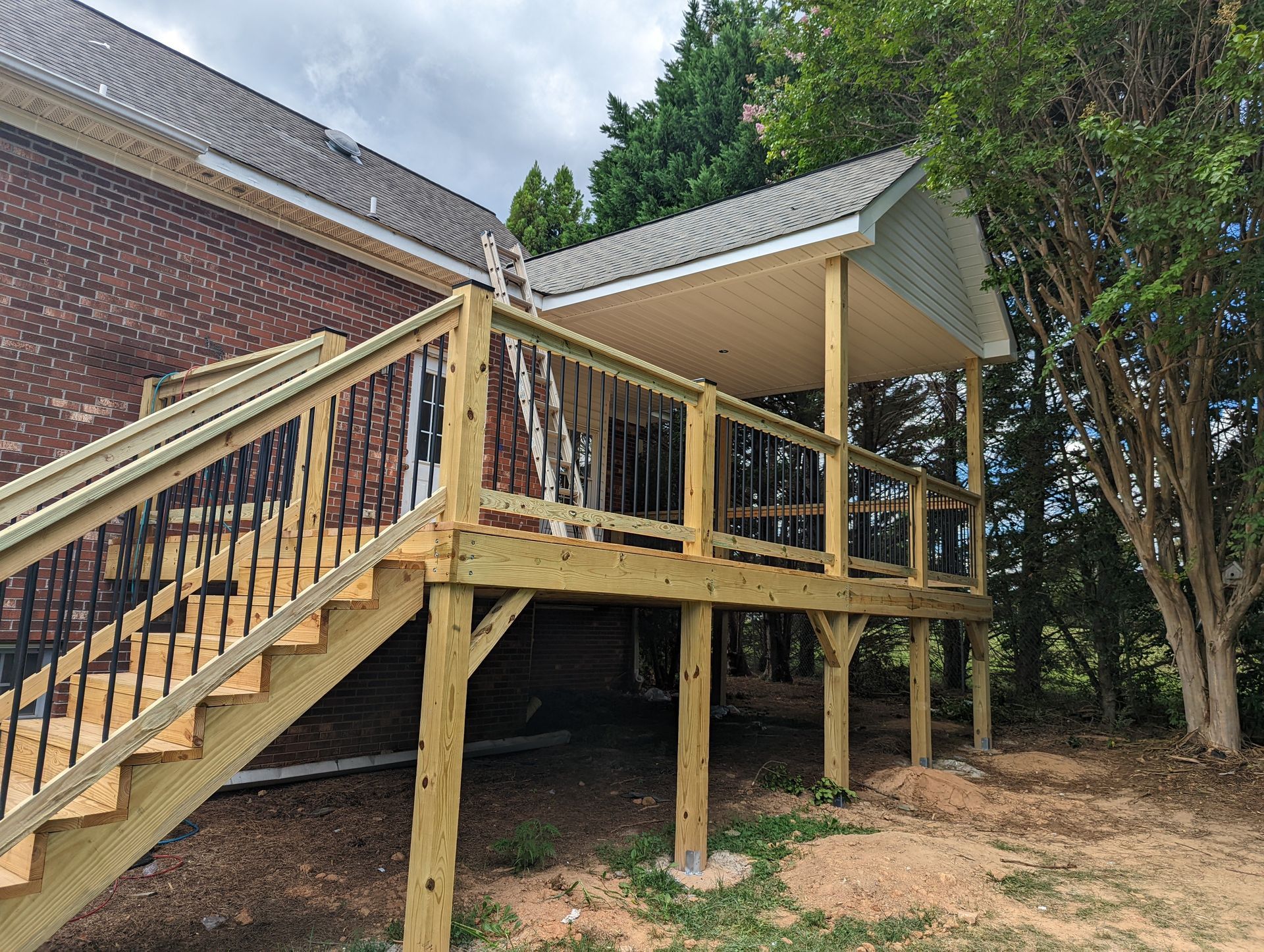 Wooden deck with stairs and a covered section attached to a brick building, set in a yard with trees.