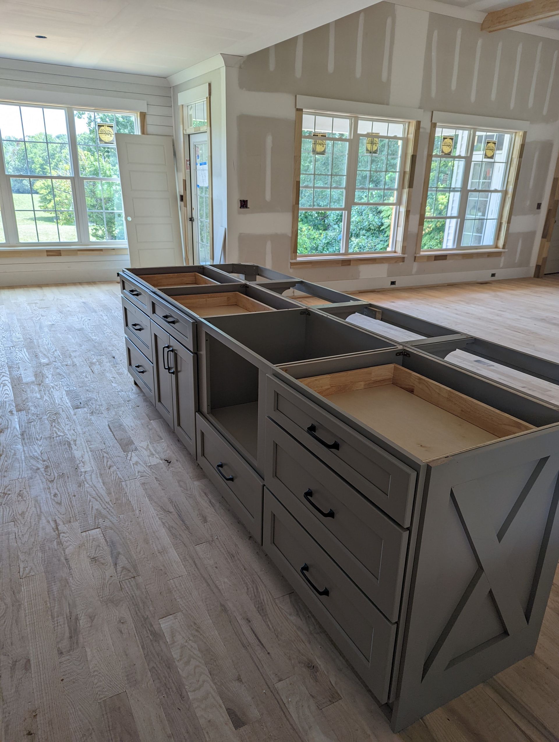 Gray kitchen island with drawers and open spaces, in a room with drywall walls and large windows.