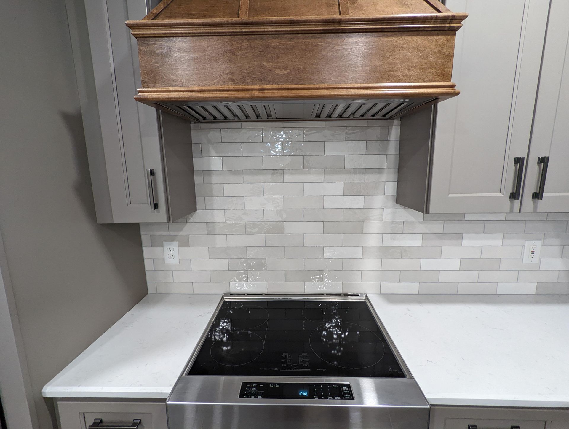 Kitchen stove with white tile backsplash, range hood, and gray cabinets. Stainless steel stove top and white countertop.