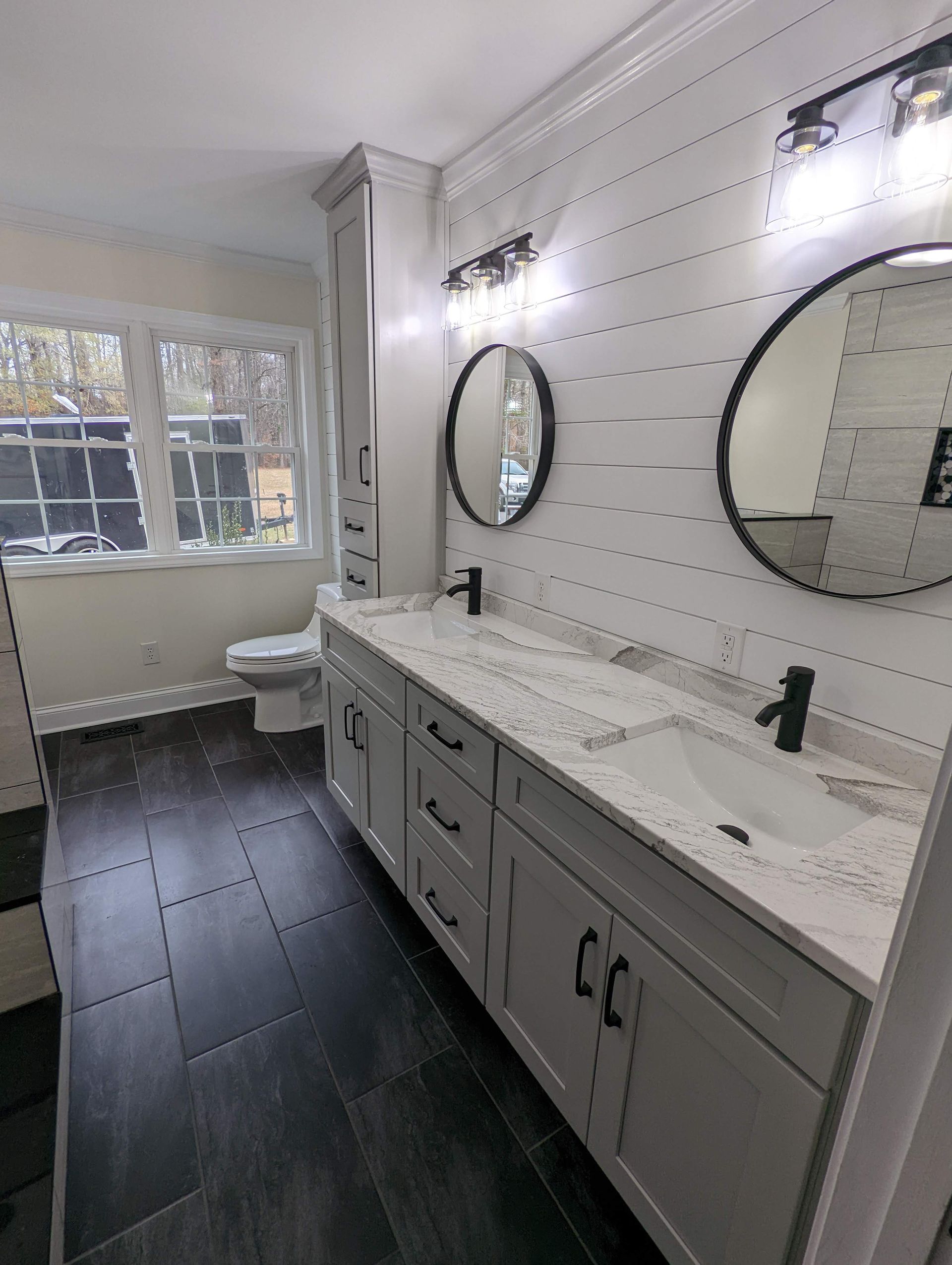 Modern bathroom with white vanity, black faucets, two round mirrors, and white shiplap walls. Black and gray tile floor.