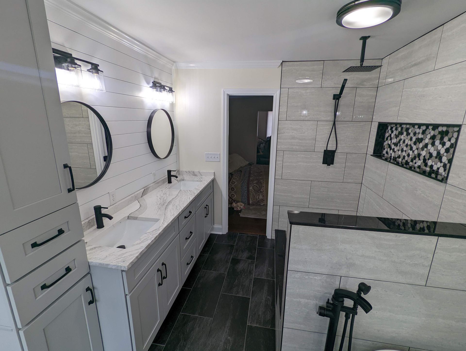 Modern bathroom with gray and white tile, black fixtures, and double vanity. A walk-in shower is to the right.
