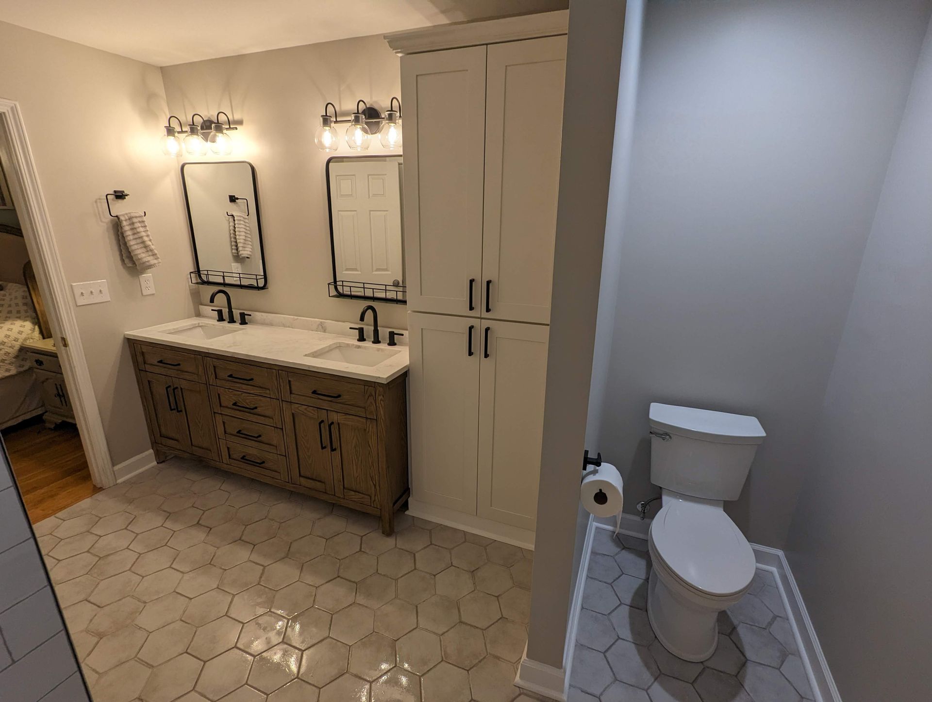 A modern bathroom with a white toilet, a double sink vanity, and a tall storage cabinet. The floor has hexagonal tiles.
