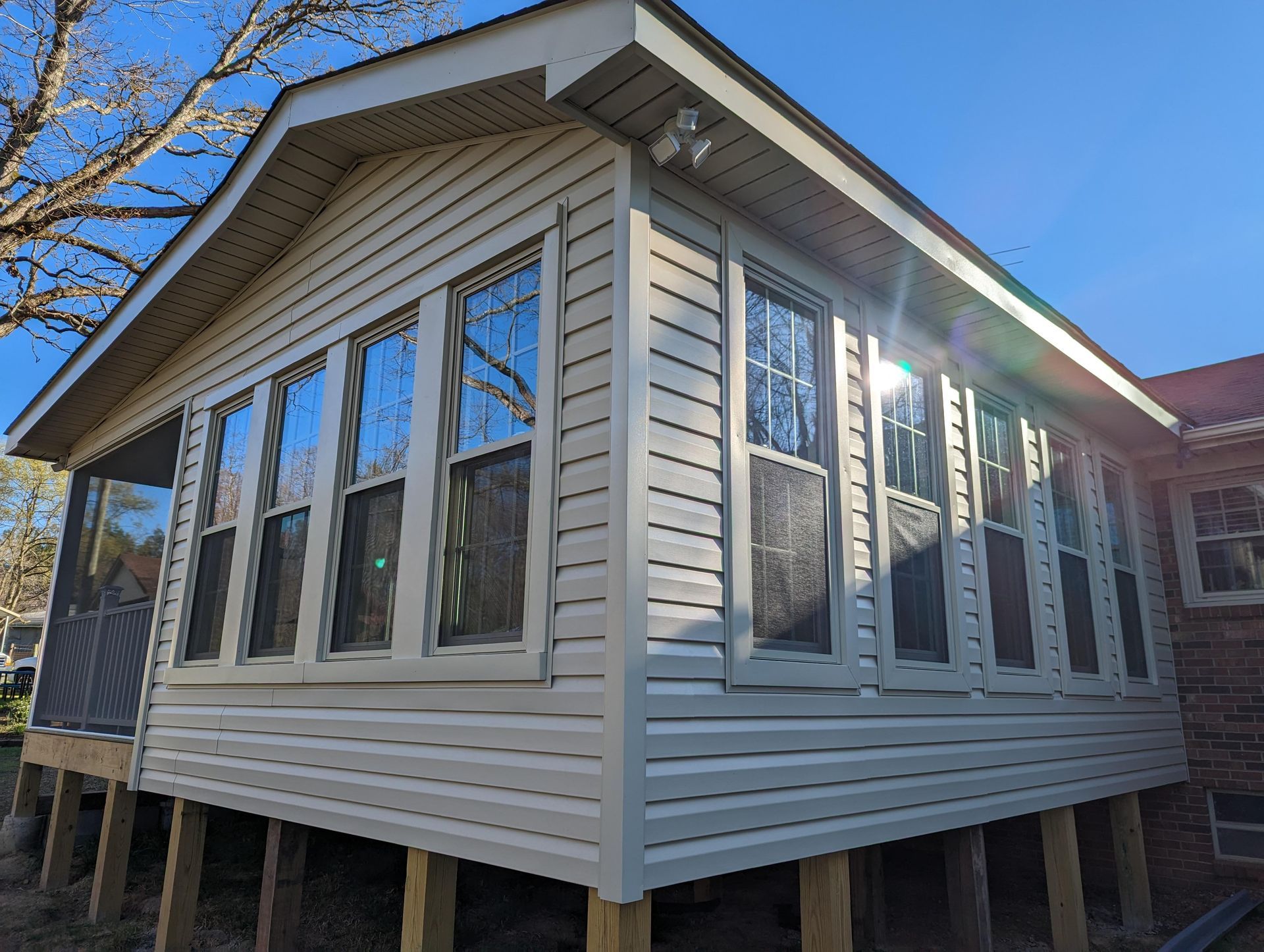 A light beige sunroom with multiple windows on a wooden frame, attached to a brick building on a sunny day.