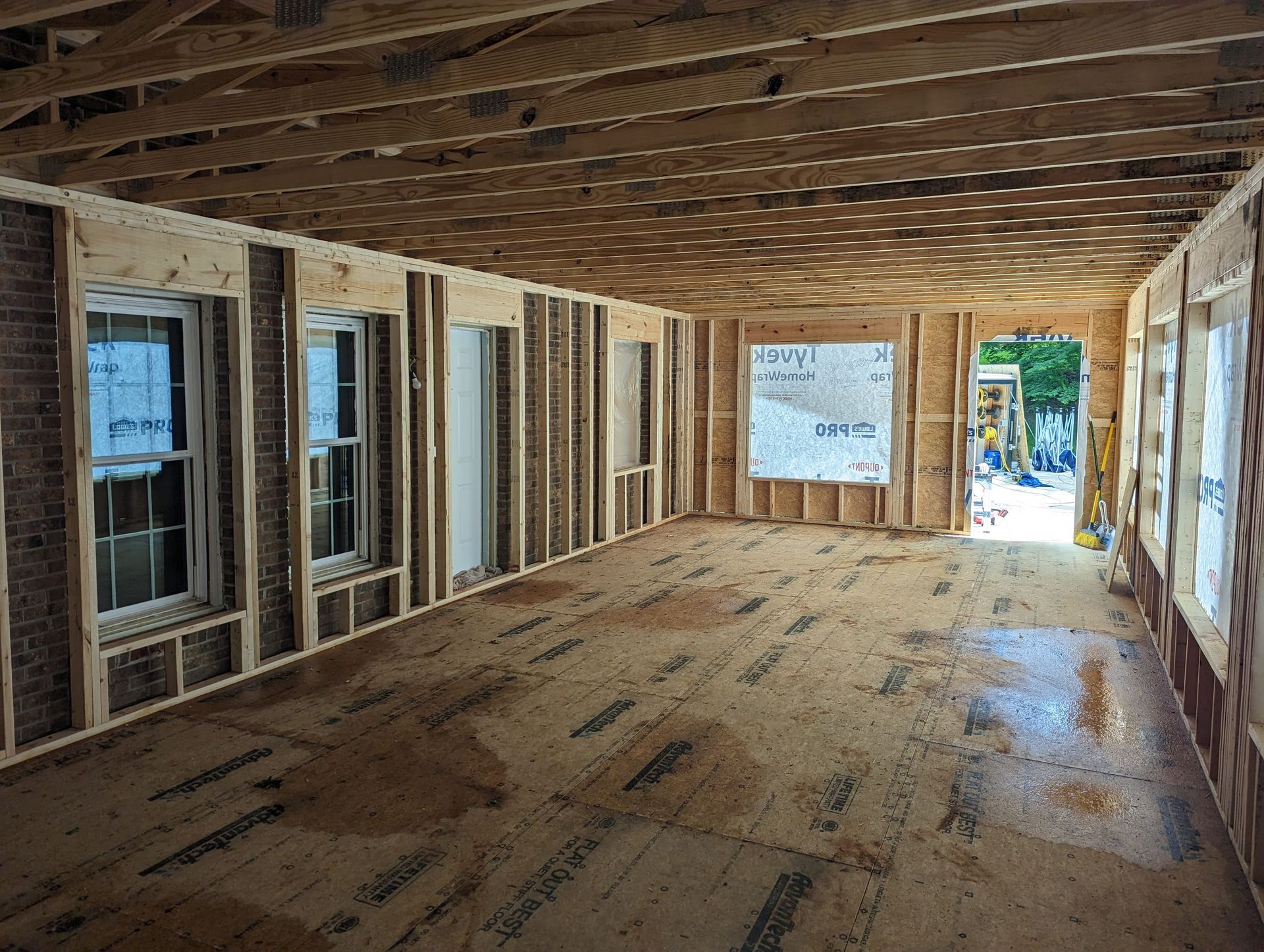 Interior view of a room under construction, with exposed wooden framing, windows, and unfinished floor.