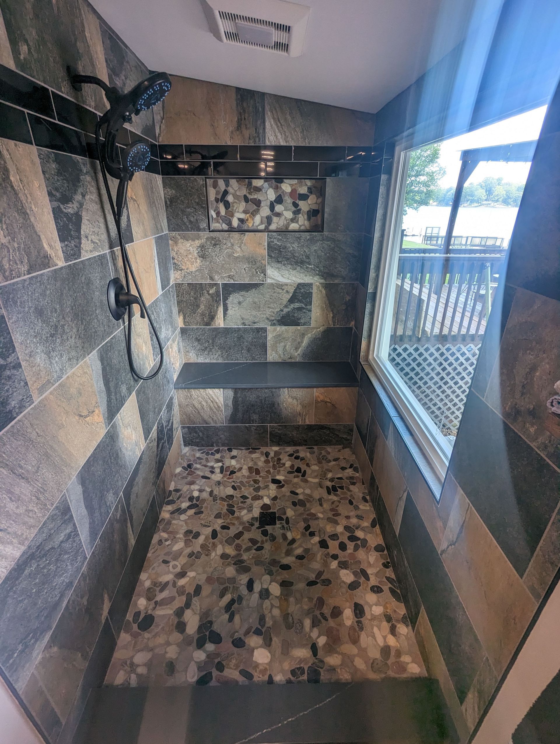 Tiled walk-in shower with pebble floor and window. Dark stone walls and showerhead.
