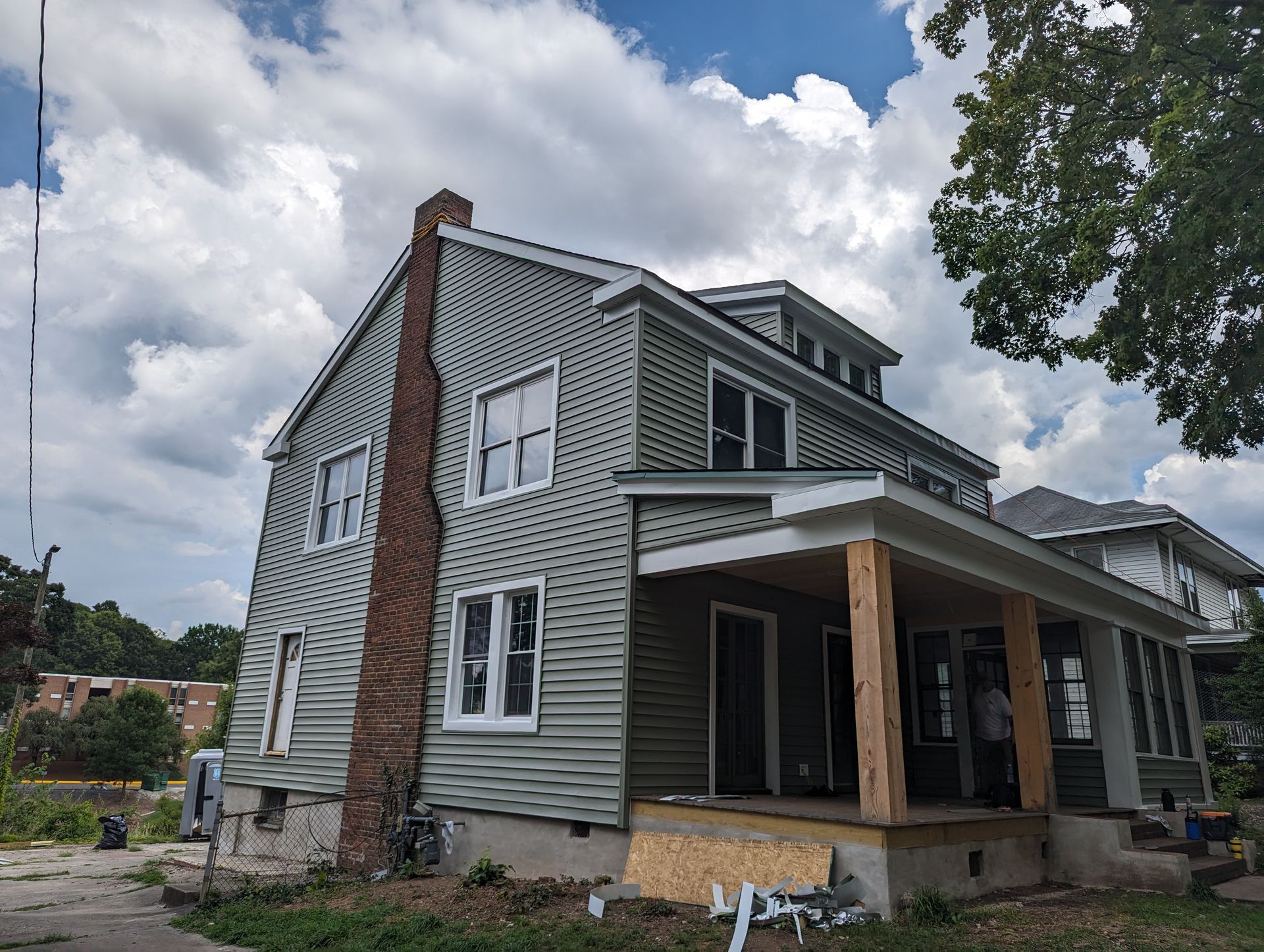 Two-story house with green siding, a brick chimney, and a partially constructed porch under a cloudy sky.