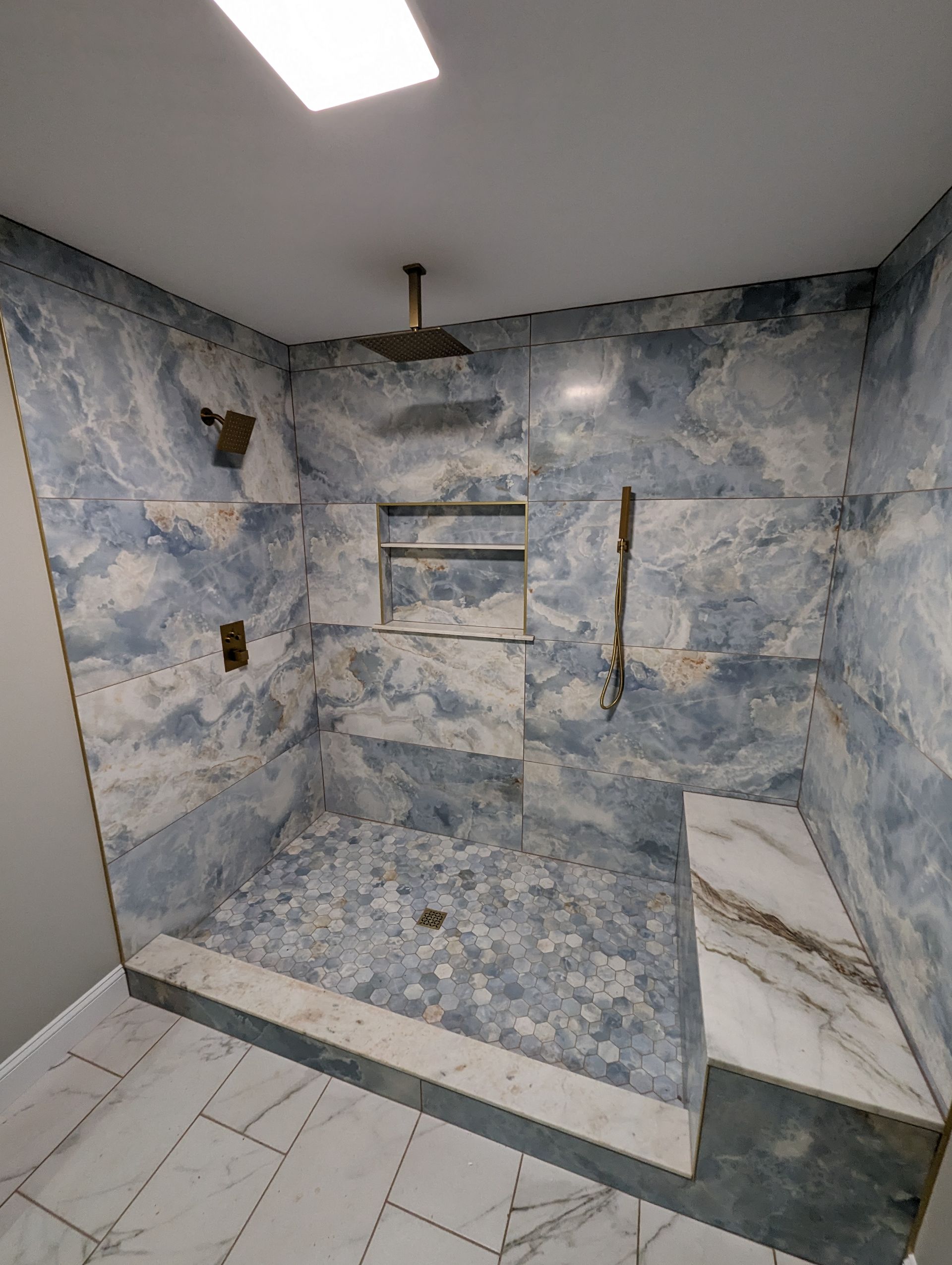 Blue and white tiled shower with a bench, shelves, and a rain shower head. Marble-look tiles line the floor.