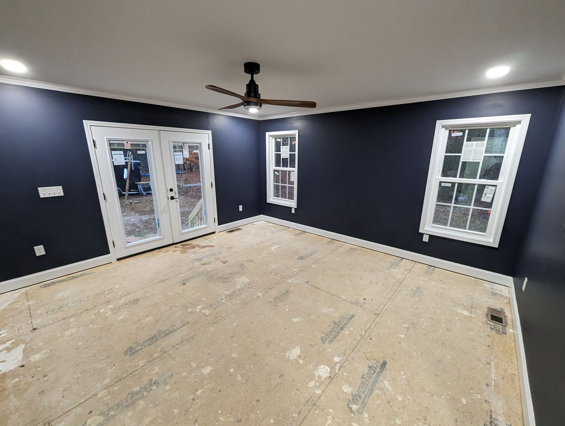 Empty room with dark blue walls, white trim, and French doors. A ceiling fan is mounted. The floor is unfinished.