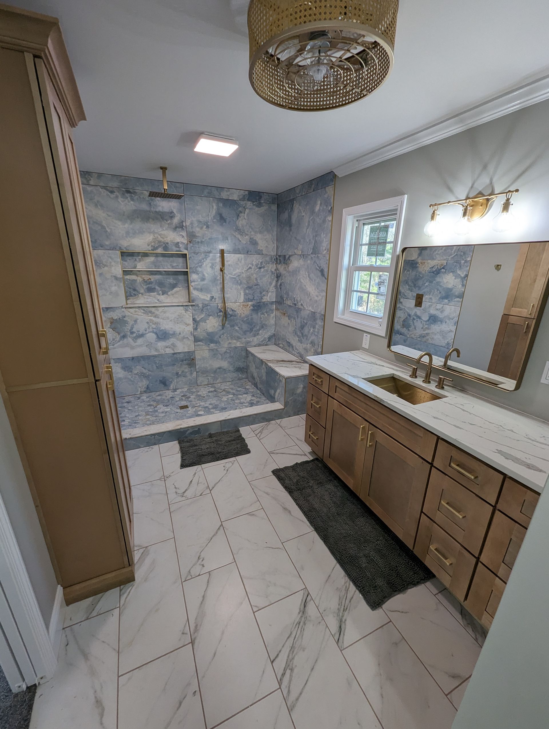Bathroom with light wood cabinets, blue tile shower, and marble-look flooring.