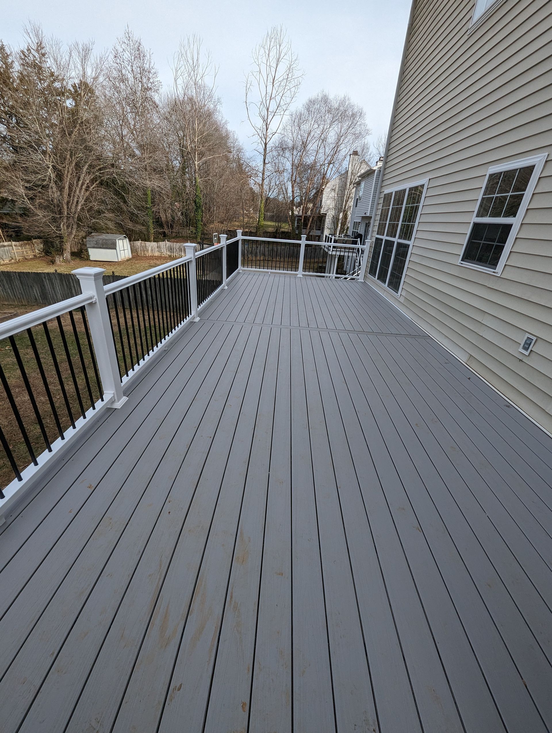 A long, gray deck with black railing and a white fence overlooking a bare tree line. The deck is attached to a beige house.