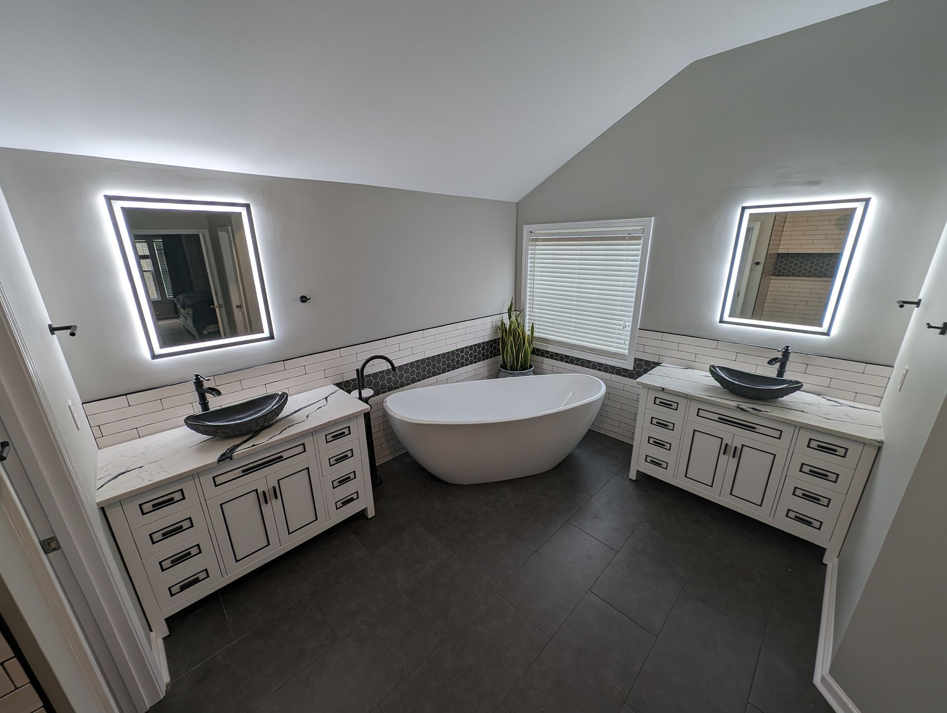 Modern bathroom with white cabinets, black sinks, and a freestanding tub. Mirrors with LED lighting flank a window.