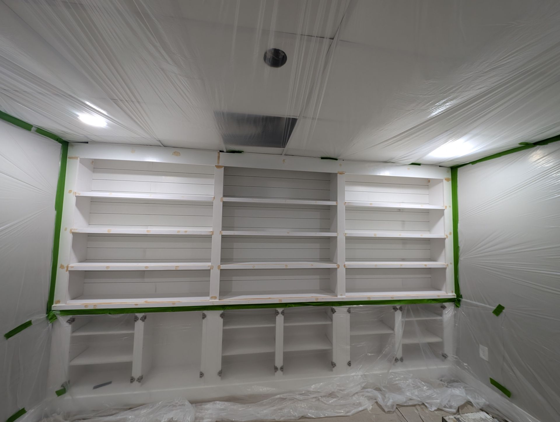 Bookshelves being painted white, covered in plastic sheeting and green painter's tape in a room.