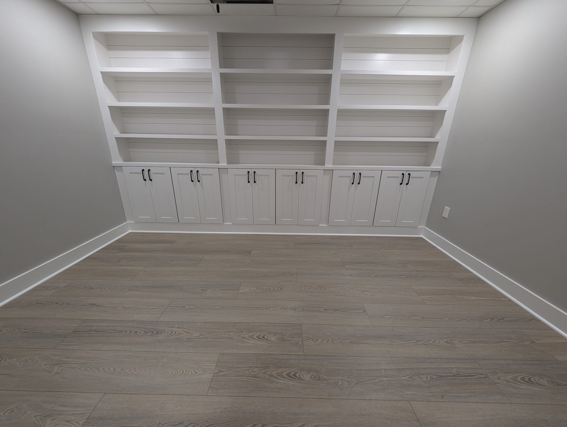 Empty room with built-in white bookshelves and cabinets on a sloped wall; gray walls, wood-look floor.