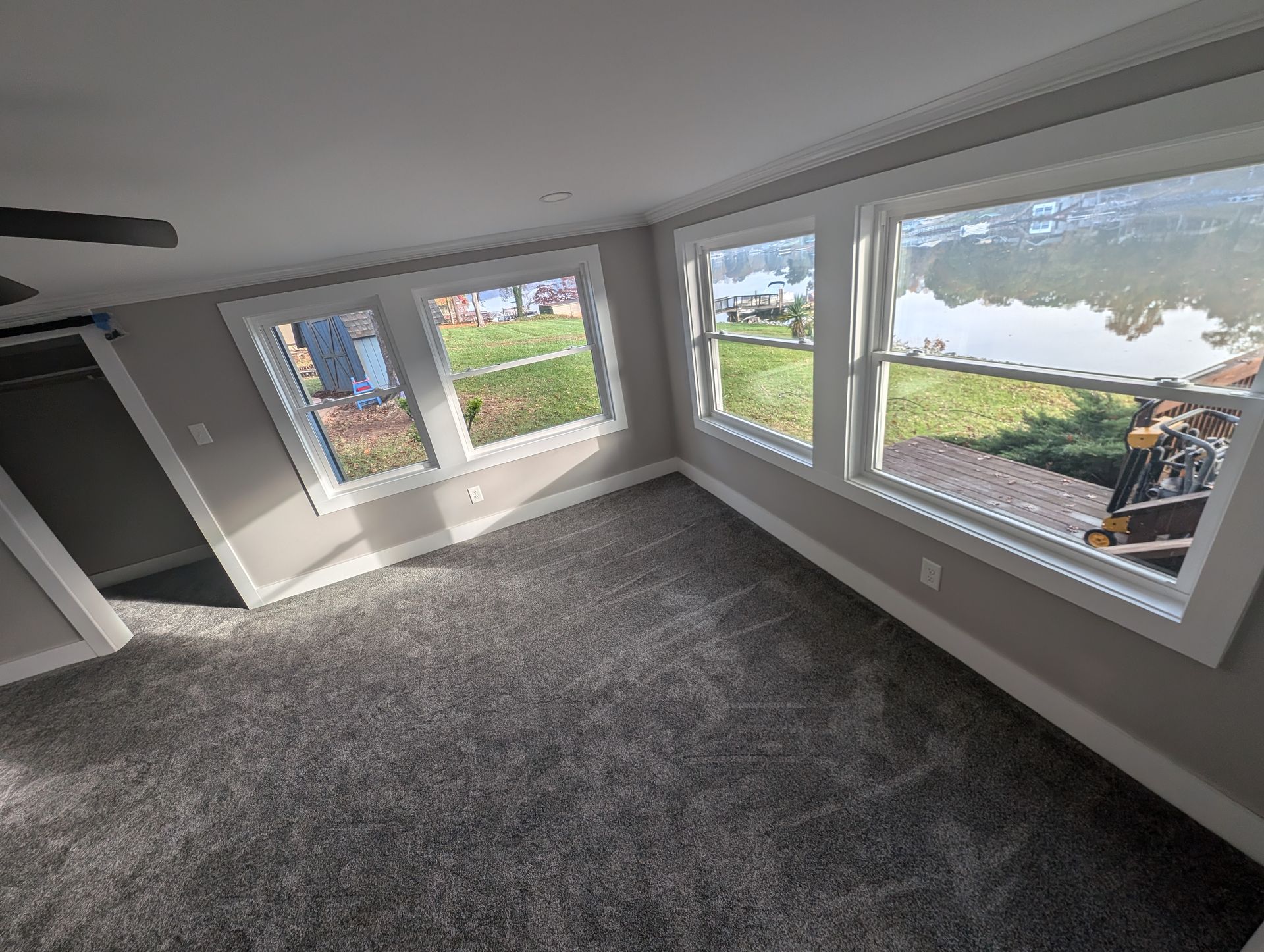 A room with gray carpet and white-framed windows overlooking a grassy yard. The room is bathed in sunlight.