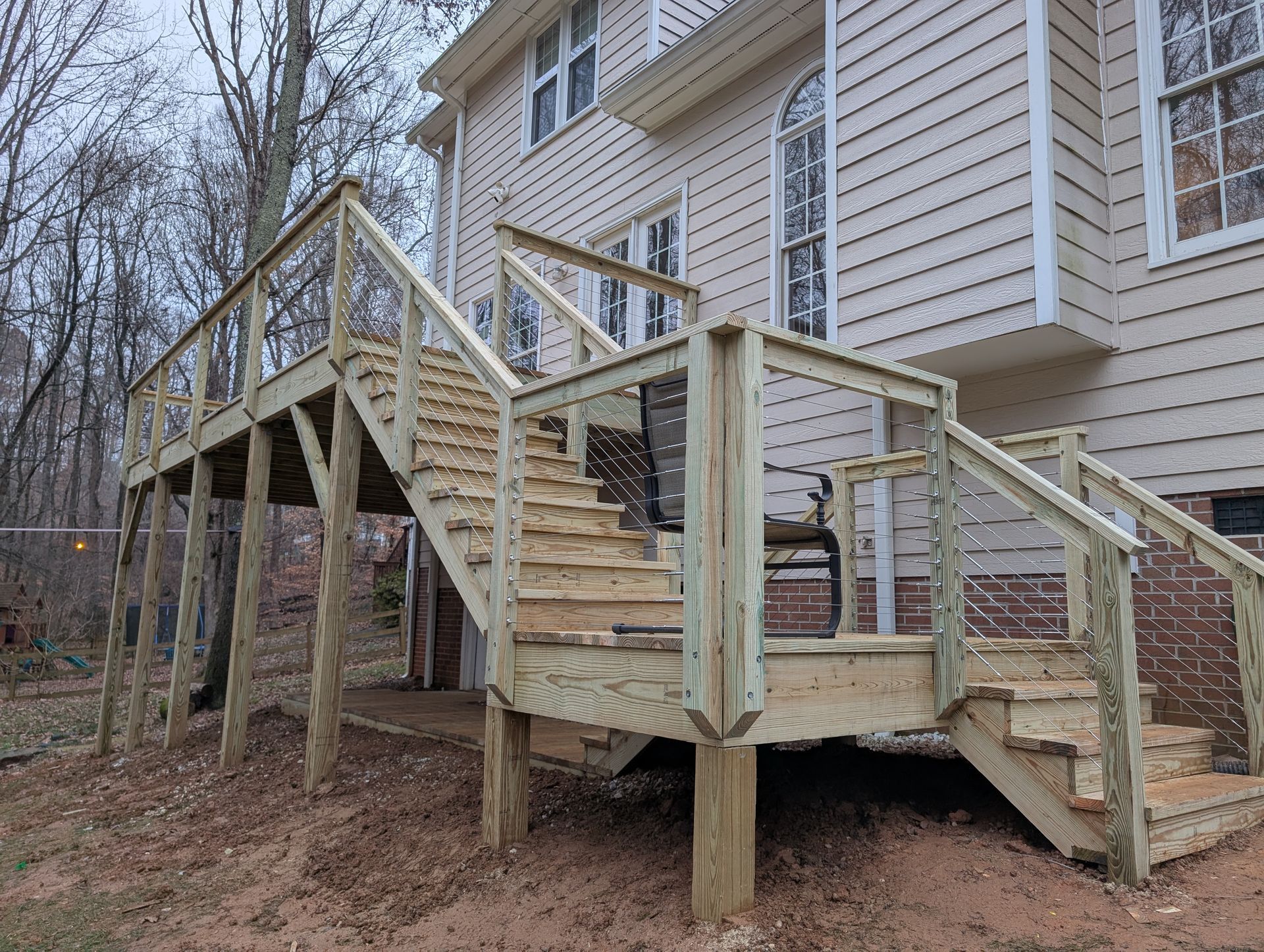 A two-story wooden deck with stairs attached to a beige house. The deck has railings and is elevated on wooden posts.