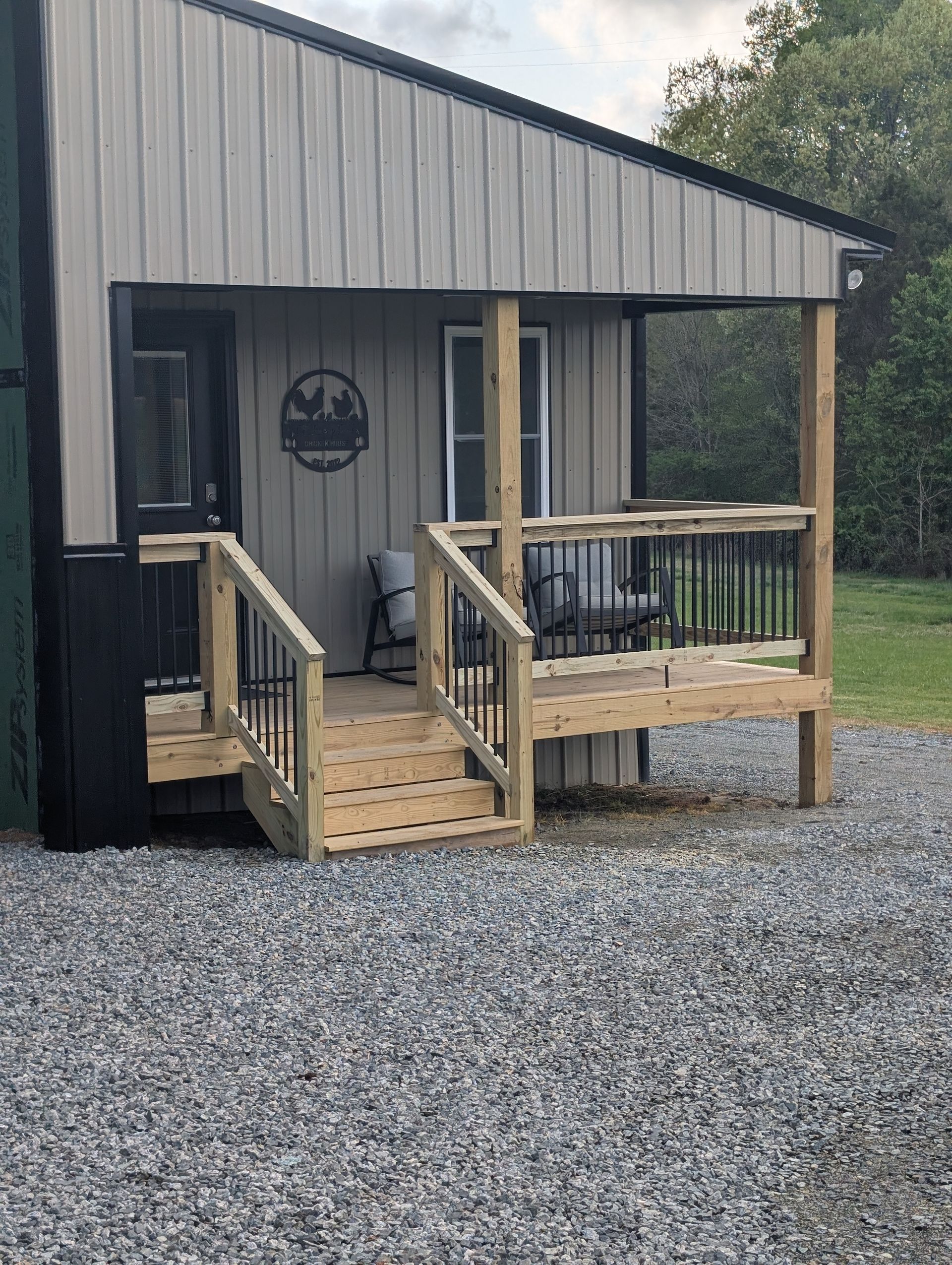 Small cabin with wooden porch and stairs. Gravel ground. Gray siding, black trim.