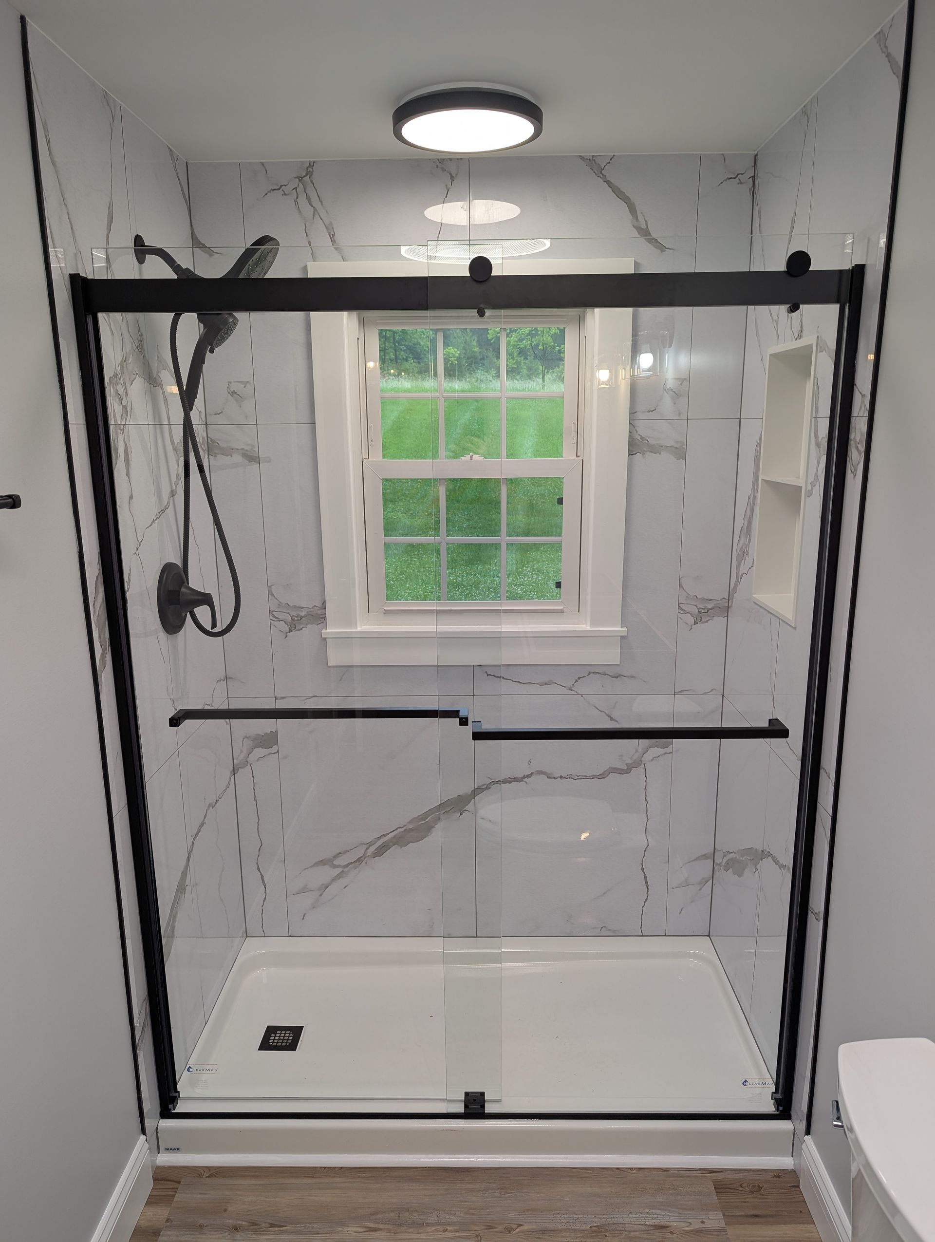 A modern shower with black framed glass doors and white marble-patterned walls. A small window is centered on the back wall.