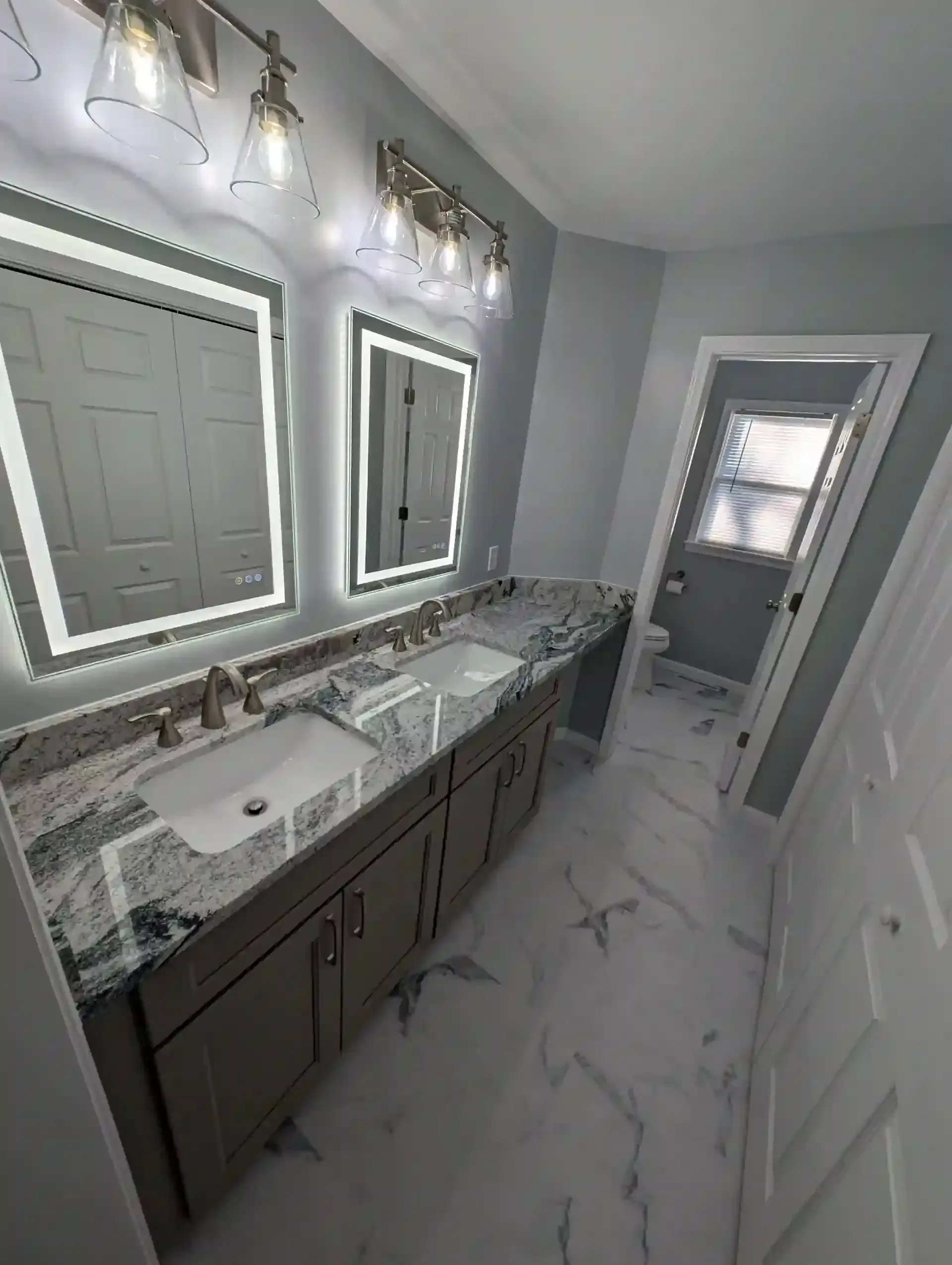 Modern bathroom with double vanity, lit mirrors, marble countertop, and a walk-in shower beyond, by N. Edwards Enterprises