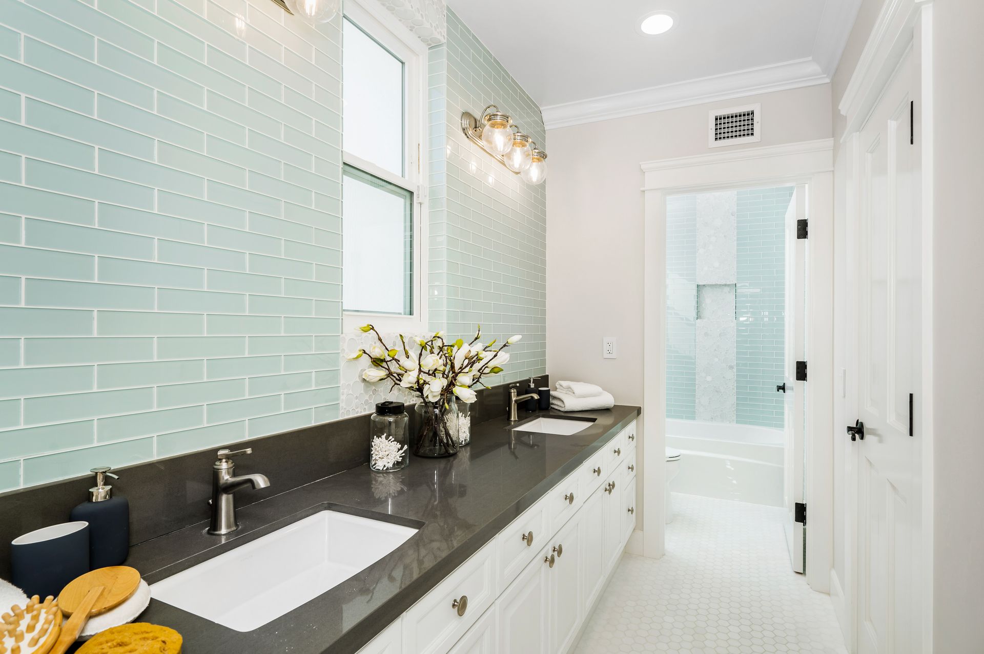 A modern bathroom with a long dark countertop, double vanity, light blue subway tile wall, and white cabinetry.