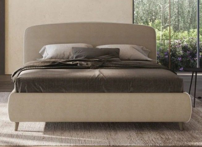 Letto moderno con testiera e pediera imbottite beige in una camera da letto illuminata da una luce soffusa.