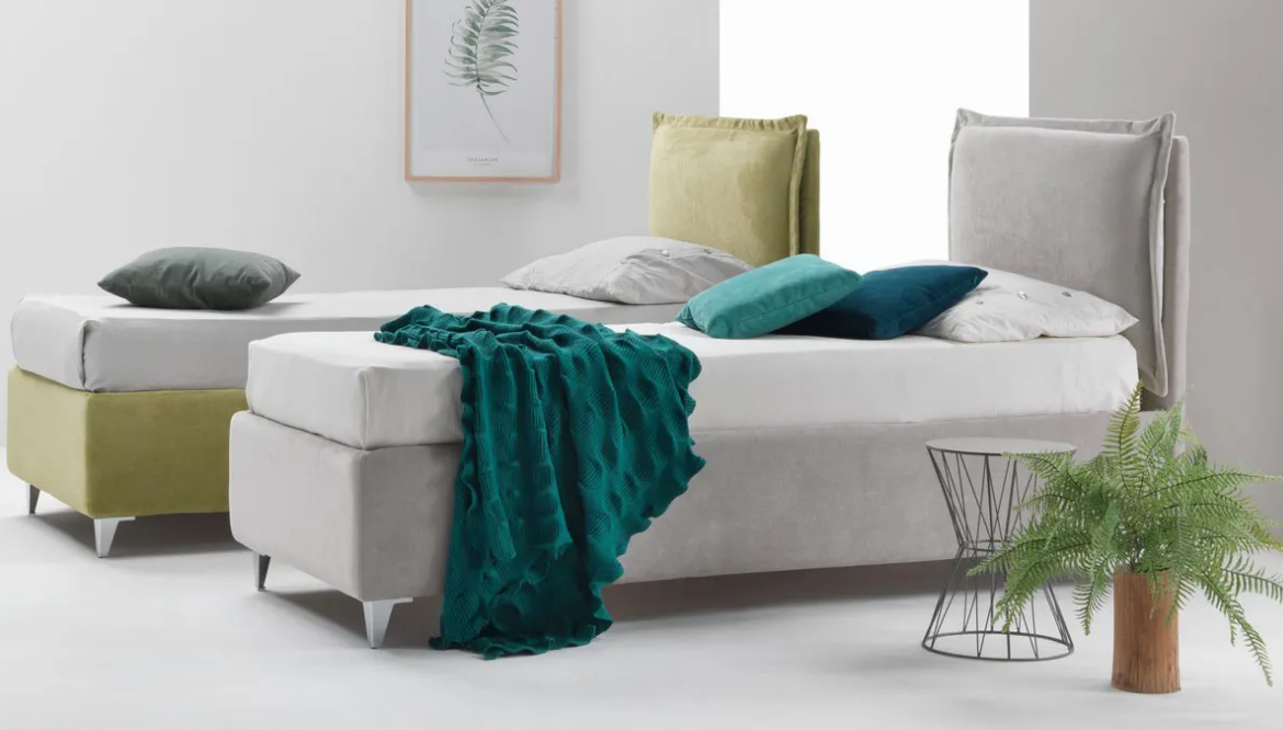 Camera da letto moderna con due letti, testiere color oliva e grigio, coperta color verde acqua e pianta in vaso vicino a un cesto di metallo.