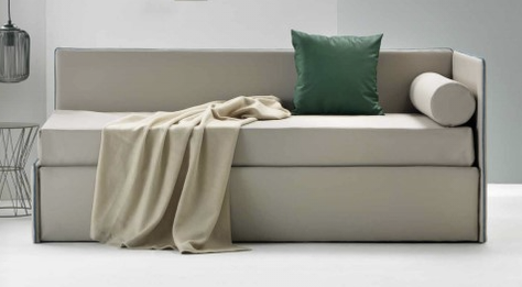 Moderno divano letto grigio con coperta beige e cuscino decorativo verde in una stanza minimalista.