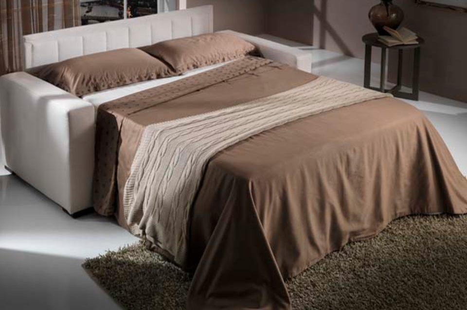Letto moderno con struttura bianca, lenzuola marroni e plaid beige in una camera da letto illuminata da una luce soffusa.