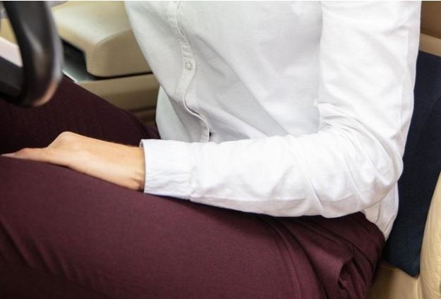 Persona seduta in auto, con indosso una camicia bianca e pantaloni bordeaux, con le mani appoggiate in grembo.