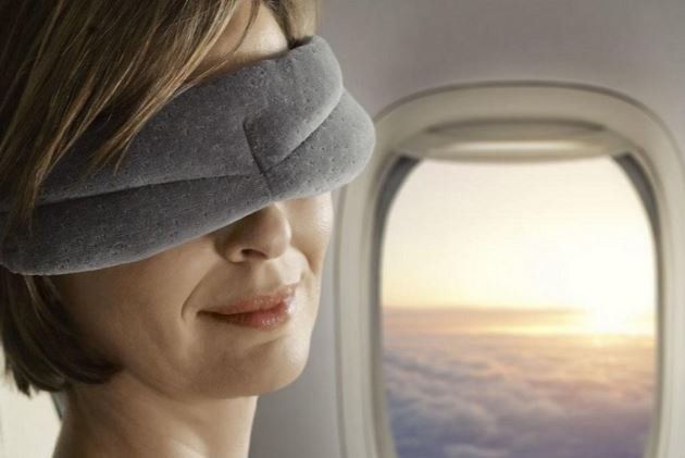 Una persona con indosso una mascherina grigia per dormire, affacciata al finestrino di un aereo al tramonto.