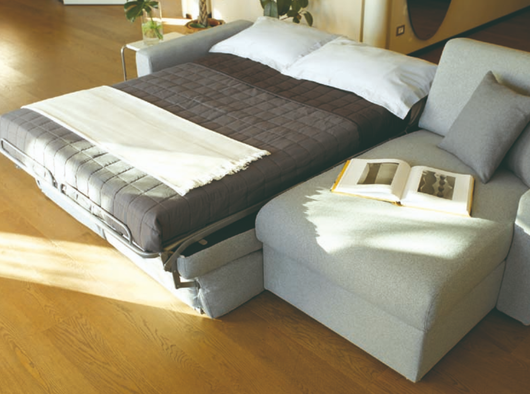 Moderno divano letto in una stanza luminosa con lenzuola grigie e un libro aperto sulla chaise longue.