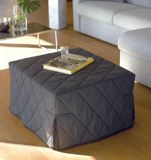 Pouf cubico trapuntato nero in soggiorno, con sopra una rivista e un bicchiere.