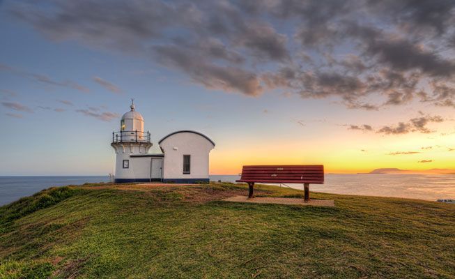 A Lighthouse and a Bench on Top of a Grassy Hill Overlooking the Ocean at Sunset — Cemak Constructions in Port Macquarie, NSW