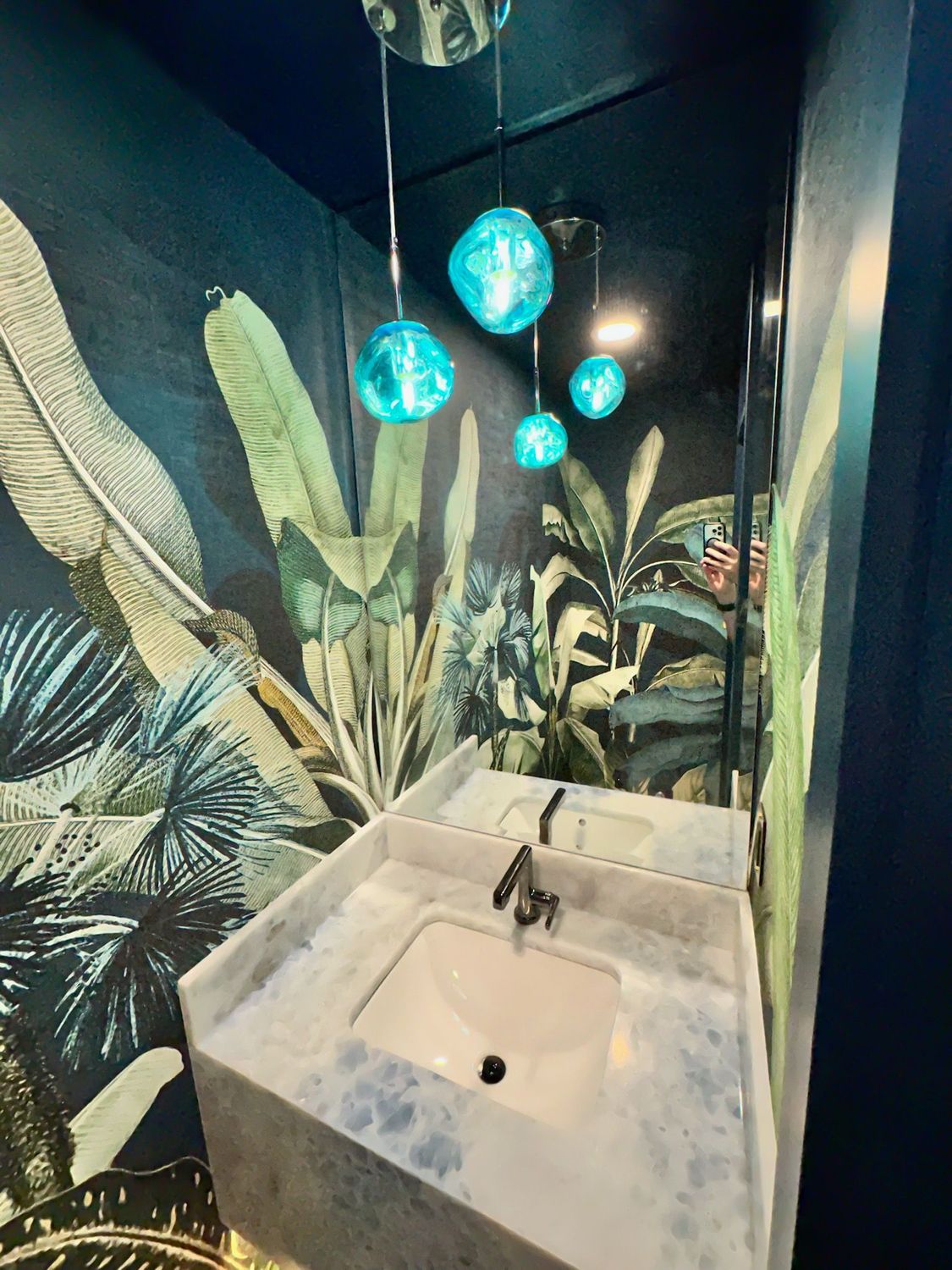 Small bathroom with tropical wallpaper, white sink, and blue pendant lights.