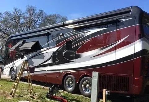 Large maroon-and-white motorcoach parked on grass under trees, side view with extended awning