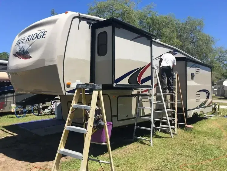 Person painting a white and gray travel trailer beside a ladder in a sunny yard