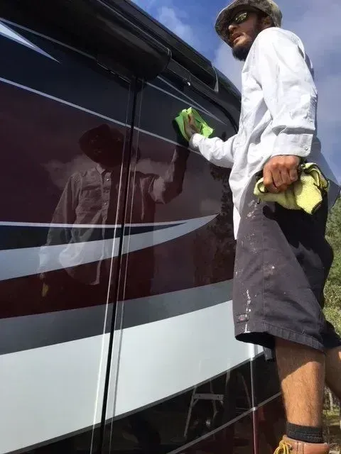 Person washing a dark maroon bus window with a green cloth outdoors.