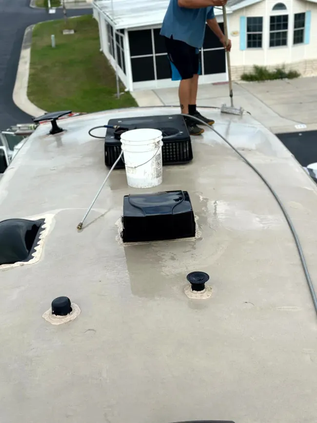 Person on a flat roof with a white bucket and black roof vents, near a house in the background