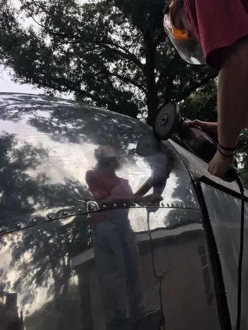 Person polishing a dark car hood outdoors, with trees reflected in the shiny surface.