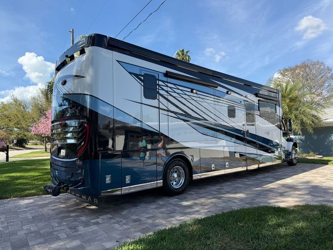 Large blue and white motorhome parked on a driveway beside a grassy yard and trees