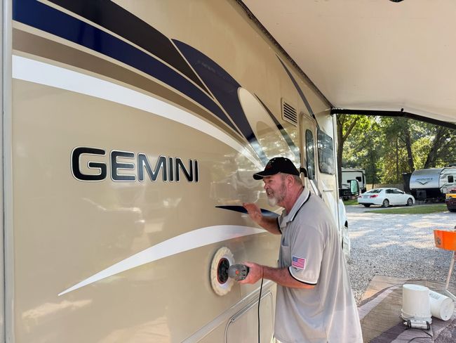 Man polishing the side of a beige Gemini RV under a canopy, with a white van and trees in the background