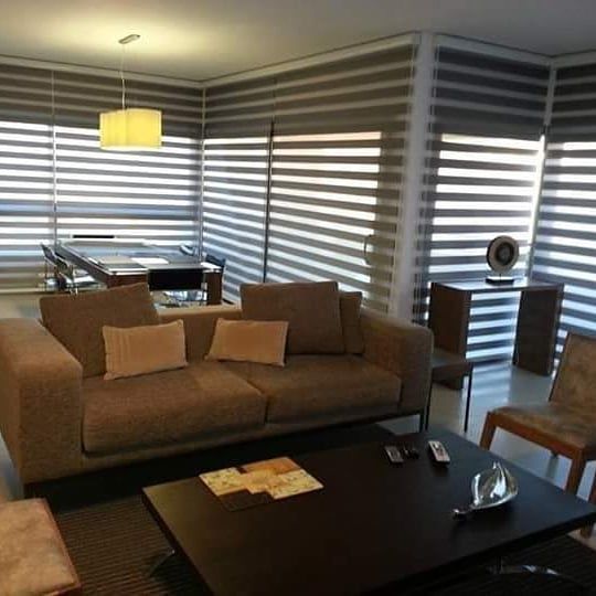 Sala de estar moderna con sofá beige, mesa de centro oscura, persianas a rayas y una pequeña lámpara de escritorio.