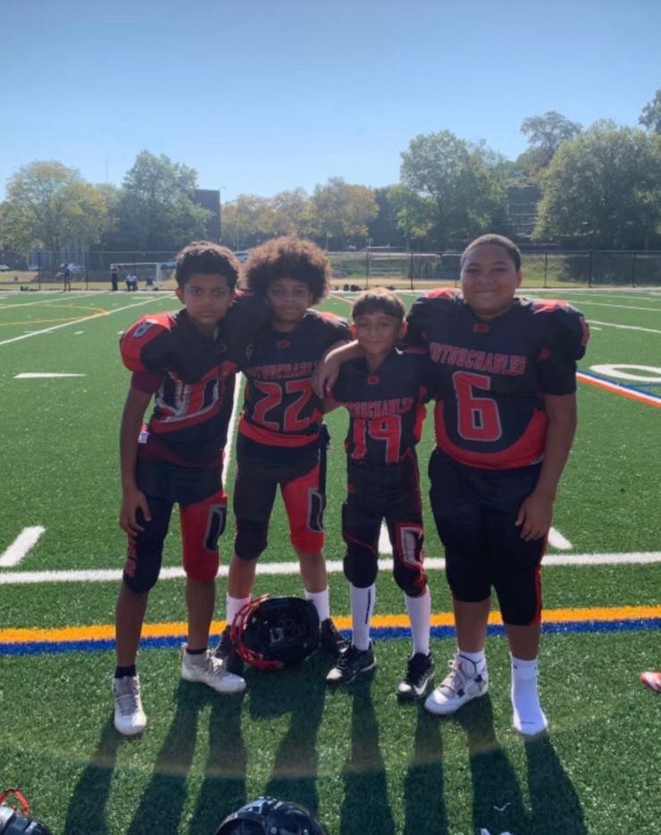 Four youth football players in black and red uniforms stand on a field with their helmets at their feet.