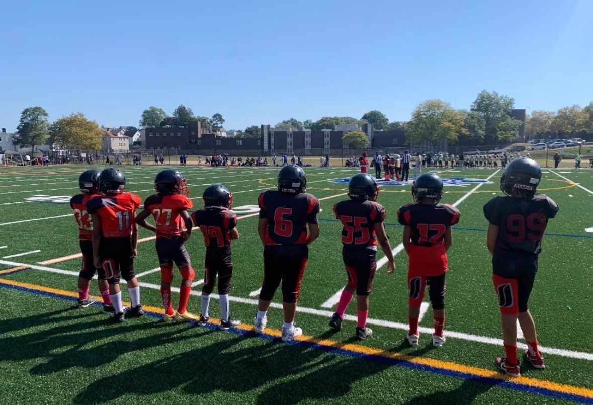 Youth football players in red and black uniforms line up on the sideline of a sunny field.