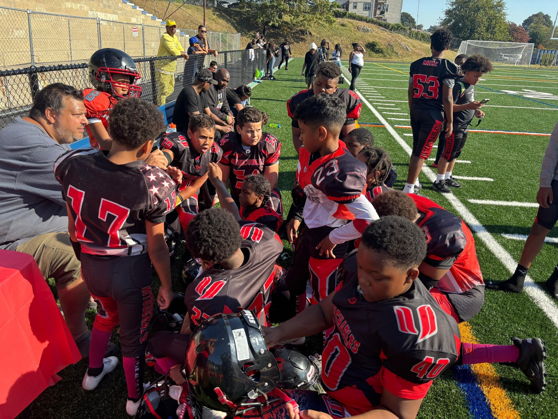 Youth football team in black and red uniforms huddles on a field during a game break.