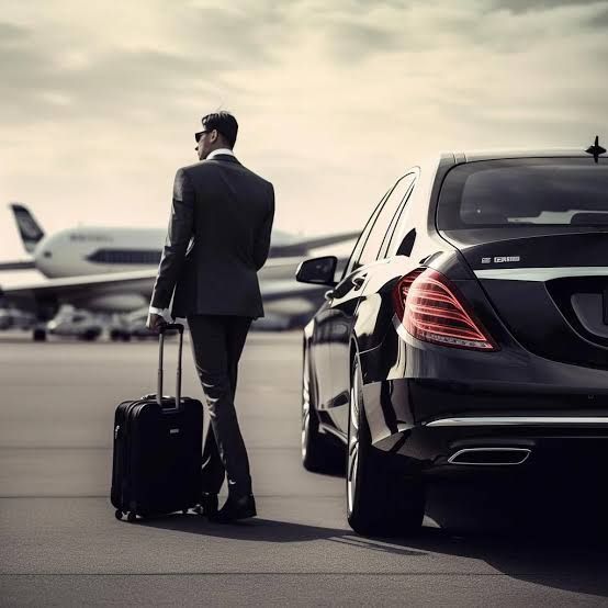 Man in a suit walking toward a black sedan at an airport, pulling a suitcase with a plane in the background.