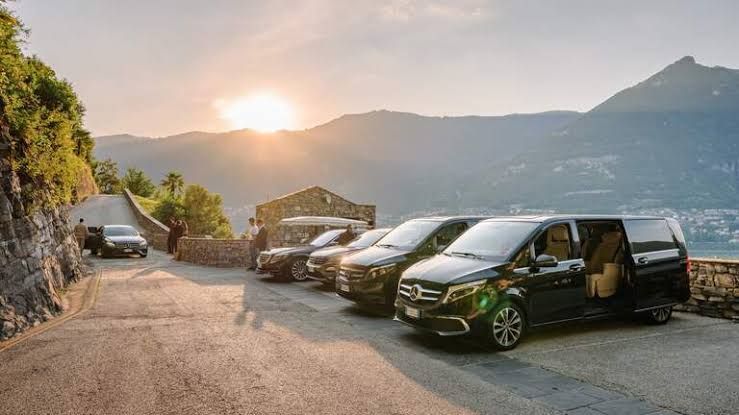 A row of black vans parked on a scenic road overlooking a lake and mountains at sunset.
