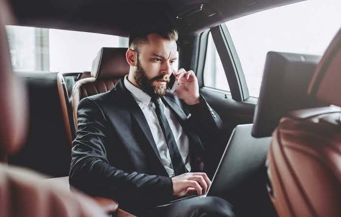 A man in a suit works on a laptop and talks on the phone in the backseat of a car.