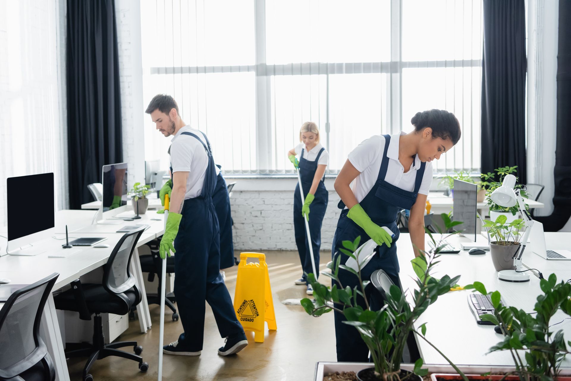 Cleaning staff vacuuming and mopping a bright office with desks and plants.
