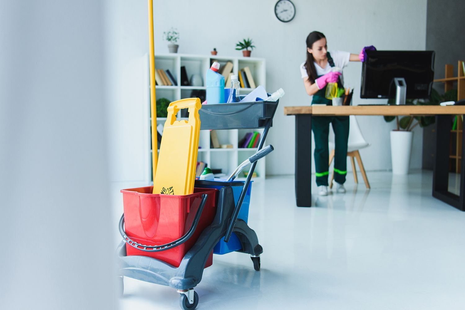 Office cleaning cart and janitorial supplies with worker wiping desk in modern workspace.